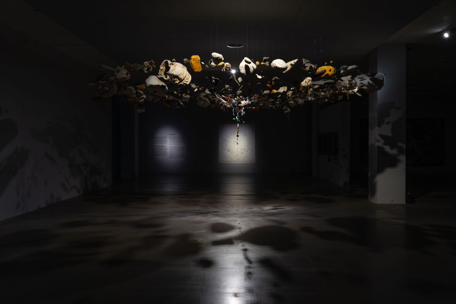 Being, 2025: a horizontal drift of plastic fragments suspended in a vast dark hall