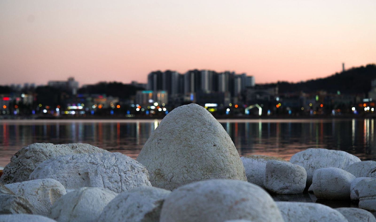 New Rocks resting at the water's edge at dusk with city lights beyond