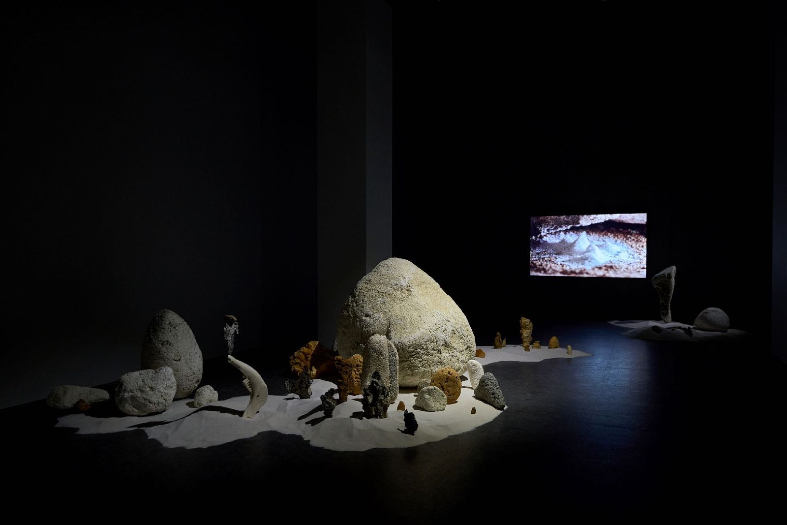 A dim gallery with pale sculptural forms and sand around a video monitor